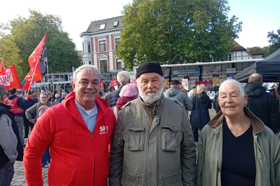 Drei lächelnde Menschen stehen nebeneinander auf dem Großflecken in Neumünster: links ein Mann mit grauem Haar in einer roten SPD-Barmstedt-Jacke, in der Mitte ein Mann mit weißem Bart und schwarzer Mütze in einer olivgrünen Jacke, rechts eine Frau mit kurzem grauem Haar in einer grünen Jacke mit Cordkragen. Hinter ihnen sieht man eine Menschenmenge, rote Fahnen, eine Bühne mit dem Banner „Für Toleranz und Demokratie!“ und eine große Linde vor einem historischen Backsteinhaus.